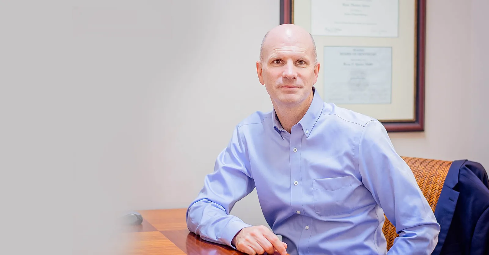 A dentist in a blue button-down shirt seated at a wooden desk in front of framed diplomas.